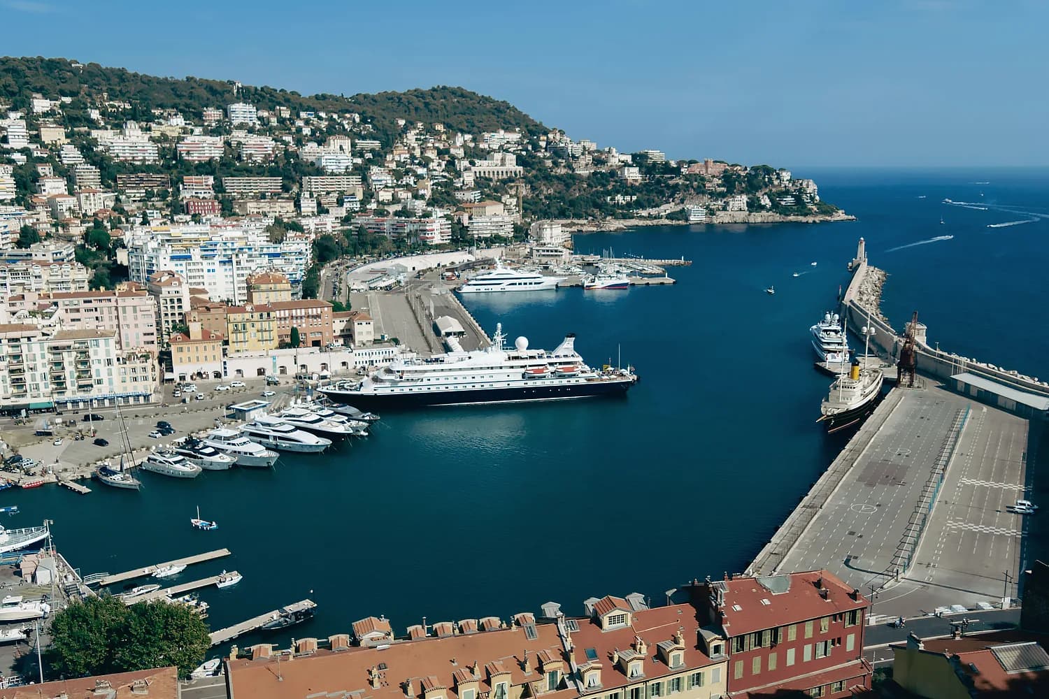 Luftaufnahme des Hafens von Nizza mit Kreuzfahrtschiffen, Jachten und Hafenbecken vor bewaldeten Hügeln