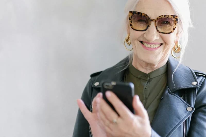 Ältere Frau mit Sonnenbrille und Lederjacke hält lächelnd ein Smartphone in der Hand und blickt aufs Display