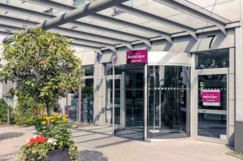 Glasvordach und drehflügeltür am Hoteleingang mit Mercure-Schildern, Topfpflanzen und gepflastertem Vorplatz.