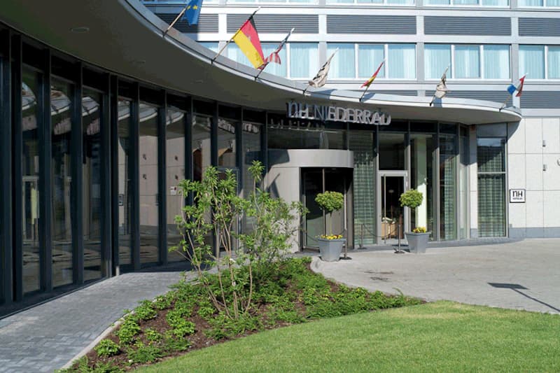 Eingangsbereich des Hotels NH Niederrad mit geschwungener Glasfassade, Drehtür, Fahnen und Vorgarten.